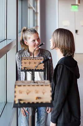 Two giggling children standing next to a Sonic Mirror instrument and speaking into its microphone.