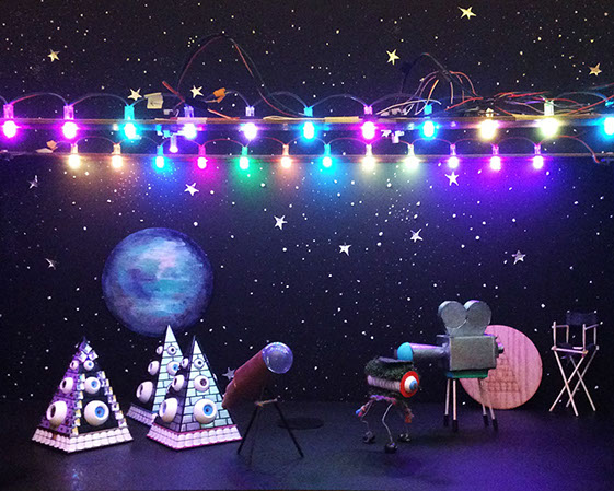 Melodyian robots on a miniature stage with an illuminated automated LED lighting system, various hand-made props, and a cosmic backdrop.